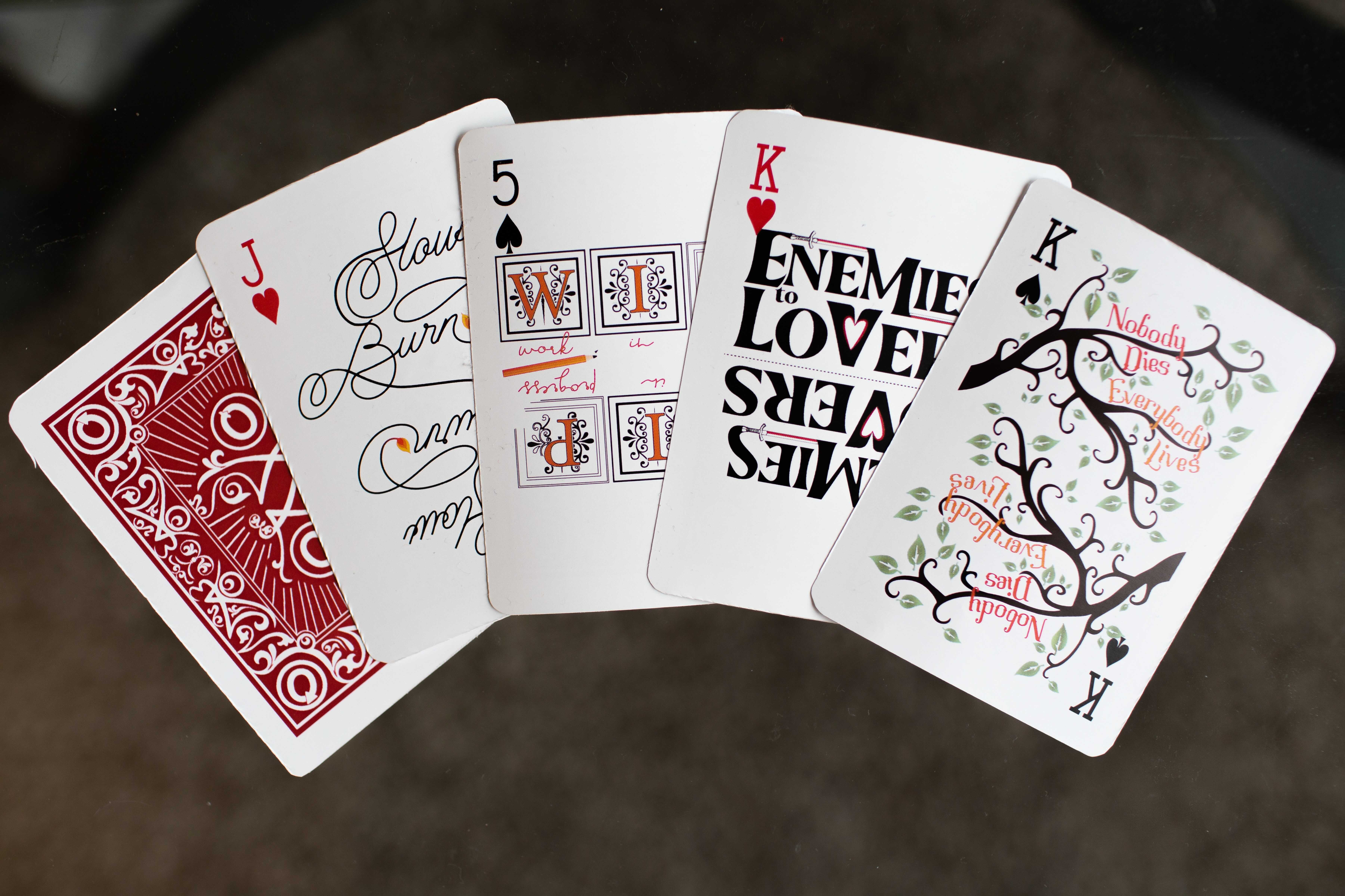 Illustrated playing cards, each representing a fannish concept: "Everybody Lives, Nobody Dies" is written on the branches of a tree, "Enemies to Lovers" is decorated with swords, the initials of "Work in Progress" are illuminated, and the last card, "Slow Burn", ends on a lit flame. A flipped-over card shows the back illustration, including the AO3 logo.