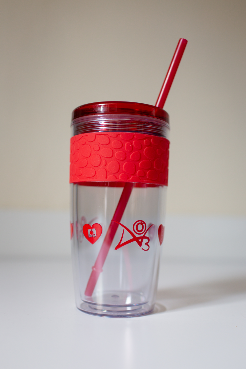 Прозрачна пластмасова чаша с червен пластмасов капак, червено уплътнение и червена пластмасова сламка. Логотата на AO3, OTW - Юридическа подкрепа, Фенлор, и Преобразуващи произведения и култури са принтирани в червено върху цилиндъра на чашата.